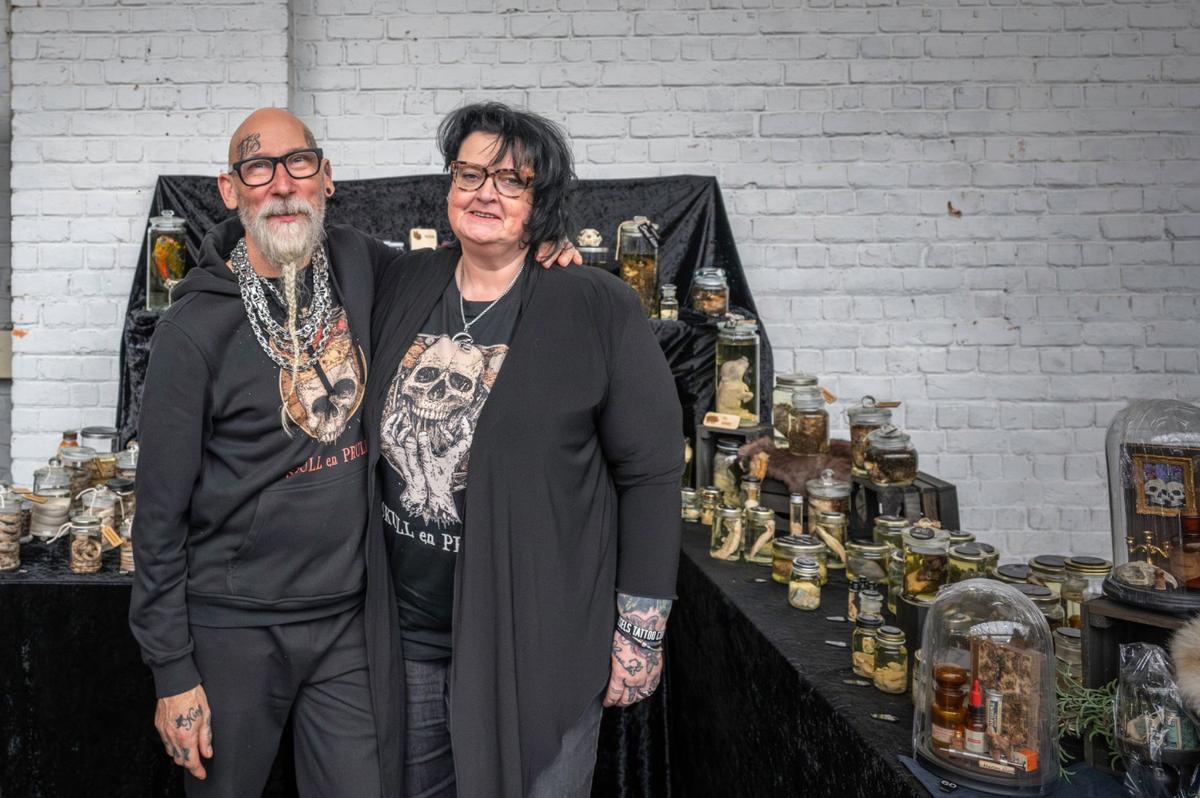 Elly and Flipke standing at their market stall in front of a white brick wall, jars and domes on the table.